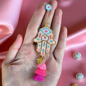 Può includere: Un ciondolo a mano di Hamsa con perline con una catena d'oro e tre nappe rosa. Il ciondolo presenta un design geometrico nei toni del rosa, blu e oro. La mano di Hamsa è tenuta in una mano su uno sfondo rosa.
