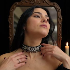 May include: A black and silver beaded choker necklace with a geometric design. The necklace has a black base with silver and clear bead accents. The choker is worn around the neck. A ring is on the finger.