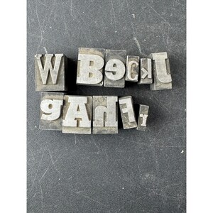 May include: A collection of eleven individual metal letterpress type blocks arranged in two rows. The blocks feature raised, reversed characters including the letters W, B, E, C, T, J, G, A, H, F, and r, showing signs of metallic wear and oxidation.