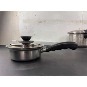 Könnte beinhalten: Ein kleiner Kochtopf aus Edelstahl mit Deckel und schwarzem Griff. Der Deckel hat einen schwarzen Knopf. Der Griff ist für einen einfachen Griff gebogen. Ein Schnellkochtopf ist im Hintergrund sichtbar.