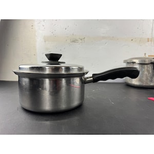 May include: A stainless steel saucepan with a black handle and lid. The pot has a shiny, reflective surface and a black knob on the lid. A second, older pot is visible in the background.