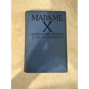 May include: A vintage book titled "MADAME X" with a blue cover. The title is in large, bold, black letters above a large "X". Below the title are the author's names, "ALEXANDRE BISSON" and "J. W. MCCONAUGHY".