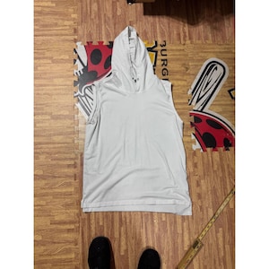 Könnte beinhalten: Ein ärmelloses weißes Kapuzenshirt mit einer Kapuze mit Kordelzug, einem geraden Saum und einer lockeren Passform. Das Kleidungsstück liegt neben einem gelben Maßband, das eine Länge von etwa 76 cm von der Schulter bis zum Saum anzeigt.