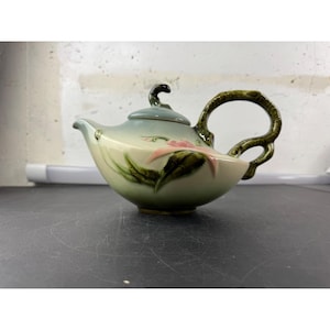 Può includere: Una teiera in ceramica con un motivo floreale. La teiera ha un gradiente verde chiaro e blu, con un fiore rosa e foglie verdi dipinte sul lato. Il manico e il beccuccio sono verde scuro.