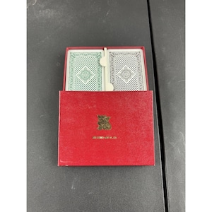 May include: A red playing card box with two decks of cards inside. The cards have green and blue backs with a repeating pattern. The box has a gold emblem and text at the bottom.