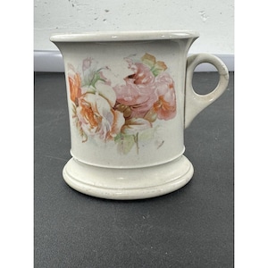 Op de afbeelding: Crèmekleurige keramische mok met een voet en een handvat. De mok heeft een bloemenontwerp met roze en oranje rozen. De mok heeft een uitlopende bovenkant en een licht gestructureerd oppervlak.