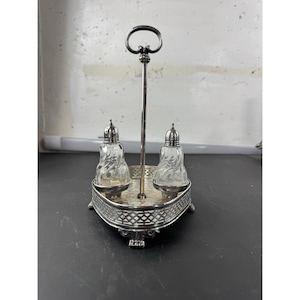 May include: Silver-plated salt and pepper shaker set. The set includes two clear glass shakers with ornate stoppers, set on a silver-toned base with a decorative handle. The base has a pierced design.