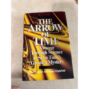 Può includere: Un libro con copertina rigida intitolato The Arrow of Time: A Voyage Through Science to Solve Time's Greatest Mystery di Peter Coveney e Roger Highfield. La copertina presenta uno sfondo scuro con motivi astratti di luce giallo oro vorticosi.