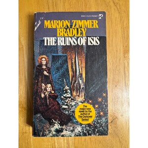 May include: A vintage paperback book titled "The Ruins of Isis" by Marion Zimmer Bradley. The cover features a dark, dramatic illustration of two women in cloaks, a tall structure, and the text "The major new novel by the author of The Darkover Series!"