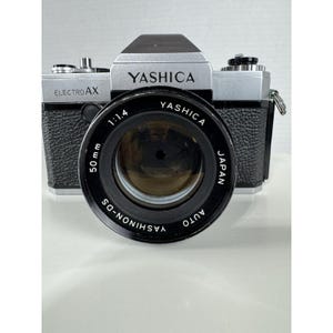 May include: A Yashica Electro AX 35mm film camera with a 50mm lens. The camera body is silver and black, with the Yashica logo prominently displayed. The lens has the markings "1:1.4" and "Yashinon-DS Auto". The camera is in good condition.