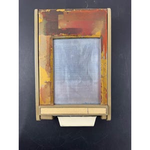 May include: A vintage rectangular picture frame with a distressed, colourful border. The frame has a central, silver-toned area, and a beige base with a white tray. The border has shades of red, yellow, and brown.
