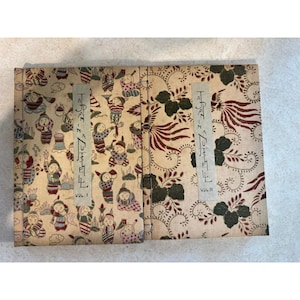 May include: Two rectangular books titled Japan in a Nutshell Vol. I and Vol. II. The first cover features a repeating pattern of small figures in colorful clothing. The second cover displays a dark red and green floral and bird motif.