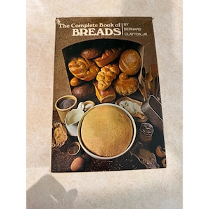 May include: A hardcover book titled The Complete Book of BREADS by Bernard Clayton, Jr. The cover features a dark brown background with an assortment of baked bread loaves, a large bowl of dough, a white pitcher, and various kitchen utensils.
