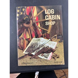 Può includere: Una copertina di catalogo vintage per il Log Cabin Shop, con un design rustico con una ruota di legno, un fucile e un cappello dell'epoca della Guerra Civile. La copertina mostra anche una spada, una pistola e un libro aperto con un'illustrazione.