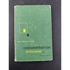 May include: A vintage book titled "Demonstration Techniques" by Mary Brown Allgood. The cover is green with a yellow and black geometric design. The words "SECOND EDITION" are printed at the bottom.