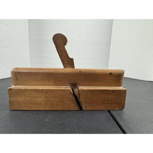 Op de afbeelding: Een vintage houten handschaaf, met een rechthoekige behuizing en een gebogen handvat. Het hout heeft een warme, natuurlijke tint, met tekenen van ouderdom en gebruik. De schaaf is ontworpen voor houtbewerking.