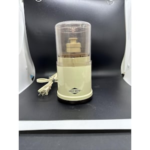 Può includere: Un macinacaffè elettrico West Bend vintage. L'apparecchio ha una base color crema con un coperchio in plastica trasparente. Il logo West Bend è visibile sulla parte anteriore. Un cavo di alimentazione si estende dalla parte posteriore. Il macinacaffè è progettato per macinare i chicchi di caffè.