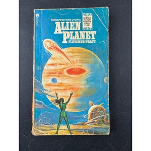 May include: Vintage paperback book titled "Alien Planet" by Fletcher Pratt. The cover features a green figure with arms raised, a planet with red and orange hues, and the text "Kidnapped into Utopia".