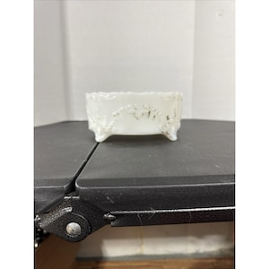 May include: A small, white, rectangular dish with ornate detailing and three small feet. The dish has a scalloped edge and a decorative pattern on the front. The dish is made of a glossy material and appears to be vintage.