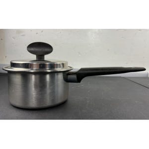 Puede incluir: Una pequeña cacerola de acero inoxidable con tapa y un mango largo negro. La olla tiene un acabado plateado y un asa negra de forma ovalada en la tapa. El mango está inclinado para facilitar su uso.