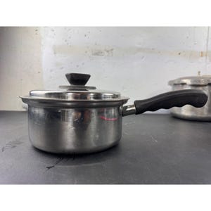 Op de afbeelding: Een kleine roestvrijstalen steelpan met een deksel en een zwarte handgreep. Het deksel heeft een zwarte knop. De steelpan staat op een donkere ondergrond. Een andere pan is zichtbaar op de achtergrond.