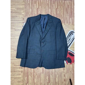 May include: A dark grey blazer with a notched lapel, two front pockets, and a two-button closure. The blazer has a dark blue interior lining and is laid flat on a patterned surface. A classic style.