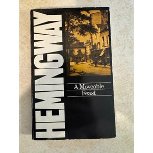 May include: A rectangular book cover with a black background and large white vertical text reading HEMINGWAY. Below this, a sepia-toned photograph depicts a street scene with buildings and people. The title A Moveable Feast is printed in white.