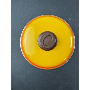Könnte beinhalten: Ein runder, gelber Glas-Topfdeckel mit einem braunen Knopf. Der Deckel hat einen silbernen Rand und eine orangefarbene Dichtung. Der Deckel liegt auf einer dunklen Oberfläche.