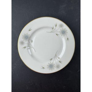 Op de afbeelding: Witte keramische plaat met een gouden rand en een bloemenontwerp met blauwe en grijze bloemen en groene bladeren. De plaat heeft een eenvoudig, elegant ontwerp, geschikt voor het serveren van voedsel.