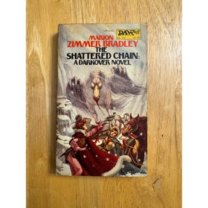 May include: Vintage paperback book titled "The Shattered Chain: A Darkover Novel" by Marion Zimmer Bradley. The cover art depicts a fantasy scene with a large creature, figures in winter clothing, and the DAW Books logo.
