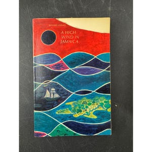 Può includere: Un libro vintage intitolato "A High Wind in Jamaica" di Richard Hughes. La copertina presenta un'illustrazione vibrante con un cielo rosso, un cerchio scuro e onde blu stilizzate. Sono raffigurati anche una barca a vela e un'isola.