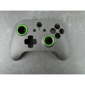 May include: A black gaming controller with green accents around the thumbsticks. The controller has a directional pad, buttons, and a central logo area. The controller is designed for video game consoles.