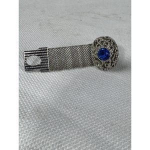 Könnte beinhalten: Silberfarbenes Mesh-Armband mit einem ovalen Mittelstück im Filigran-Stil. Das Mittelstück hat einen leuchtend blauen Edelstein. Das Armband hat einen rechteckigen Verschluss mit einer kreisförmigen Öffnung. Der Schmuck wird auf einer strukturierten, hellgrauen Oberfläche präsentiert.