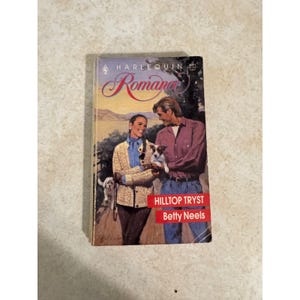 May include: A vintage paperback book titled "Hilltop Tryst" by Betty Neels. The cover shows a couple with a small dog, set against a landscape. The book is part of the Harlequin Romance series.