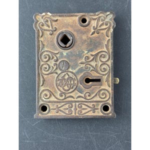 May include: Antique bronze-colored door lock plate with ornate scrollwork and a keyhole. The rectangular plate features a diamond-shaped opening, a circular element with a logo, and screw holes for mounting. A small, brass-colored lever is visible.