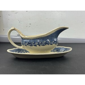 Puede incluir: Salsera de color crema con un estampado floral azul y platillo a juego. La salsera tiene un asa curva y un pico. El patrón azul se repite en el borde del platillo.