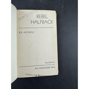 Puede incluir: Libro abierto con el título "REBEL HALFBACK" de Joe Archibald. La portada es de color blanquecino con texto negro. Publicado por The Westminster Press, Filadelfia. Texto manuscrito visible en la esquina superior izquierda.