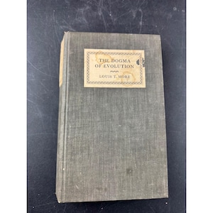 Może przedstawiać: Zabytkowa książka zatytułowana "The Dogma of Evolution" autorstwa Louisa T. More'a. Książka ma wyblakłą zieloną okładkę z prostokątną etykietą z tytułem i nazwiskiem autora. Krawędzie książki noszą ślady zużycia.