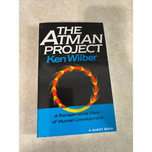 May include: A paperback book titled The Atman Project by Ken Wilber. The cover is black with white and blue text, featuring a circular graphic of orange and yellow arrows. The bottom section is blue with the text A Transpersonal View of Human Development.