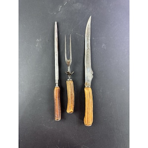 May include: A vintage carving set featuring a sharpening steel, carving fork, and carving knife. The handles are made of a light brown material, possibly antler, and the blades are silver. The set is displayed against a dark background.