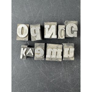 May include: A collection of nine individual metal letterpress blocks arranged in two rows. The blocks feature raised, reversed characters including O, P, N, C, G, K, A, E, M, and U, designed for traditional printing processes with a metallic silver finish.
