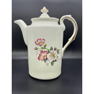 Op de afbeelding: Crèmekleurige keramische theepot met een bloemenontwerp met roze en witte bloemen en groene bladeren. De theepot heeft een goudkleurige handgreep en deksel, en een tuit. Een klassiek stuk voor het serveren van thee.