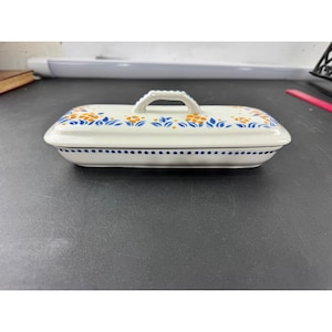 May include: A white ceramic soap dish with a lid. The dish is rectangular with rounded edges and a small handle on the lid. It is decorated with blue and orange floral patterns and a blue dotted border.