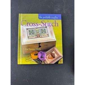 Puede incluir: Un libro titulado "Cross-Stitch" de Liz Turner Diehl, parte de la serie "the portable crafter". La portada presenta un diseño de punto de cruz en una caja de madera, con carretes de hilo y tijeras.