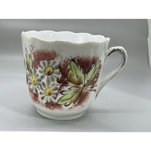 May include: White teacup with a scalloped rim and a floral design. The cup features a handle and is decorated with daisies in shades of yellow, green, and brown. The background has a brown and white mottled pattern.