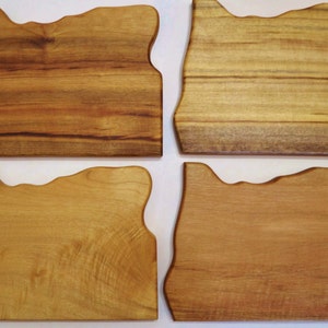 May include: Four wooden cutting boards, each shaped like the state of Oregon. The boards vary in color from light to dark brown, showcasing the natural wood grain. The boards are displayed on a white surface.