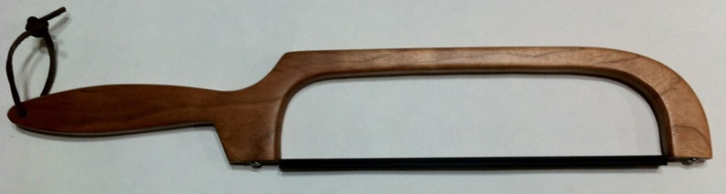 May include: A wooden-handled saw with a black metal blade. The handle is made of light brown wood and has a leather loop at the end.