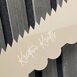 May include: A beige cake decorating tool with a scalloped edge and a serrated edge. The text "Krestive Krafts" is written in a cursive font on the tool. The background is a dark gray, textured surface.