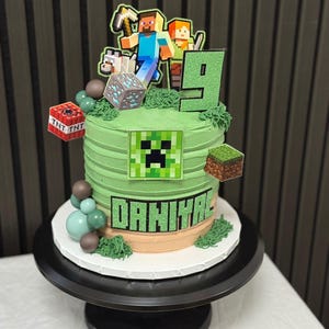Può includere: Una torta di compleanno verde a due livelli decorata con elementi a tema Minecraft. La torta presenta una faccia di Creeper, personaggi pixelati e il nome "DAHIYA". Ulteriori decorazioni includono un numero 9, blocchi TNT e sfere verdi e marroni.