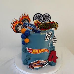 May include: A blue frosted cake decorated with Hot Wheels themed toppers. The cake features a Hot Wheels logo, race car images, flames, and the number 4. The name "Yahya" is also displayed on the cake.